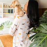 Back side of model wearing the Long-Sleeved Women's Pajama Set in Dachshund while holding a blonde dachshund with a large green plant 