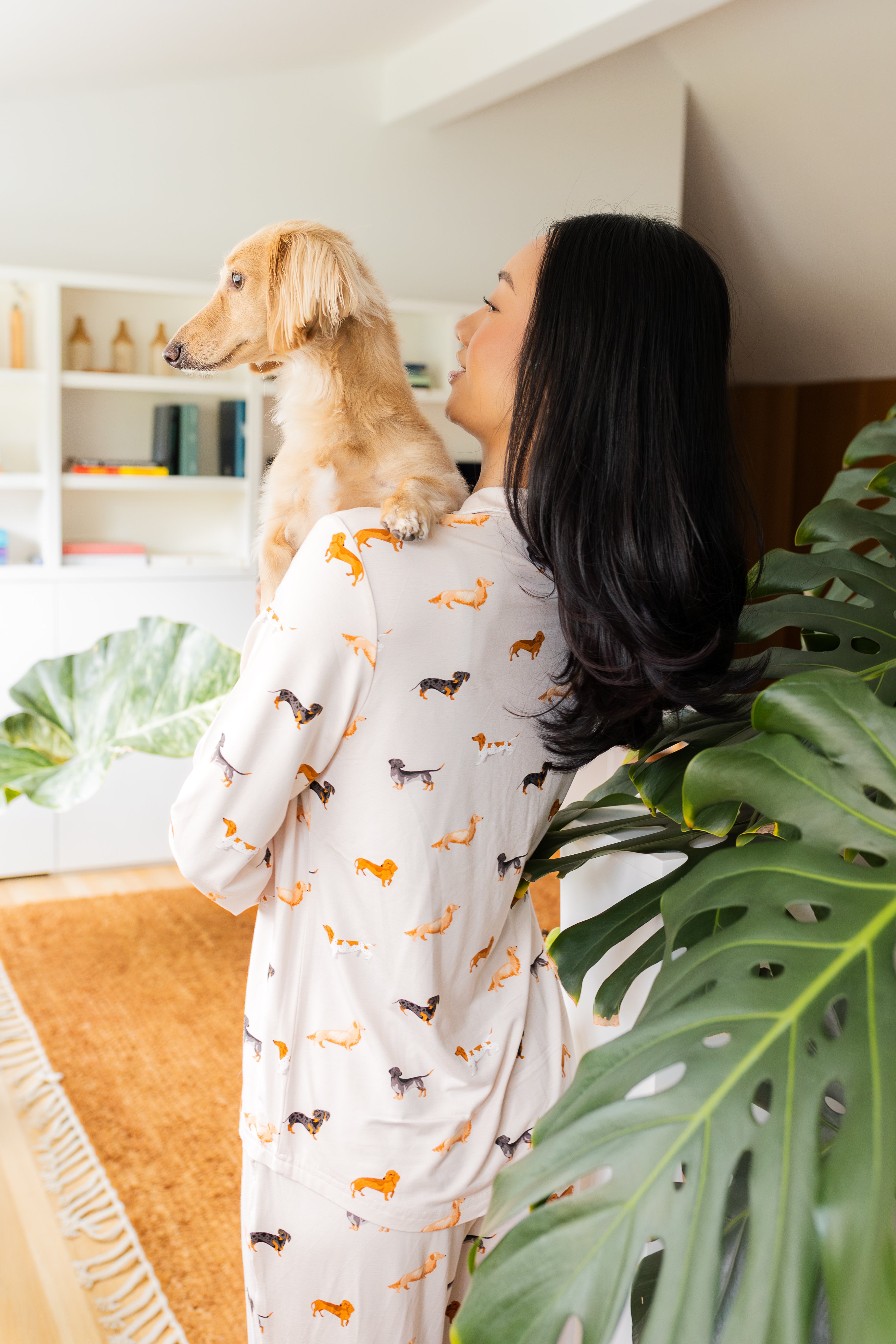 Back side of model wearing the Long-Sleeved Women's Pajama Set in Dachshund while holding a blonde dachshund with a large green plant 