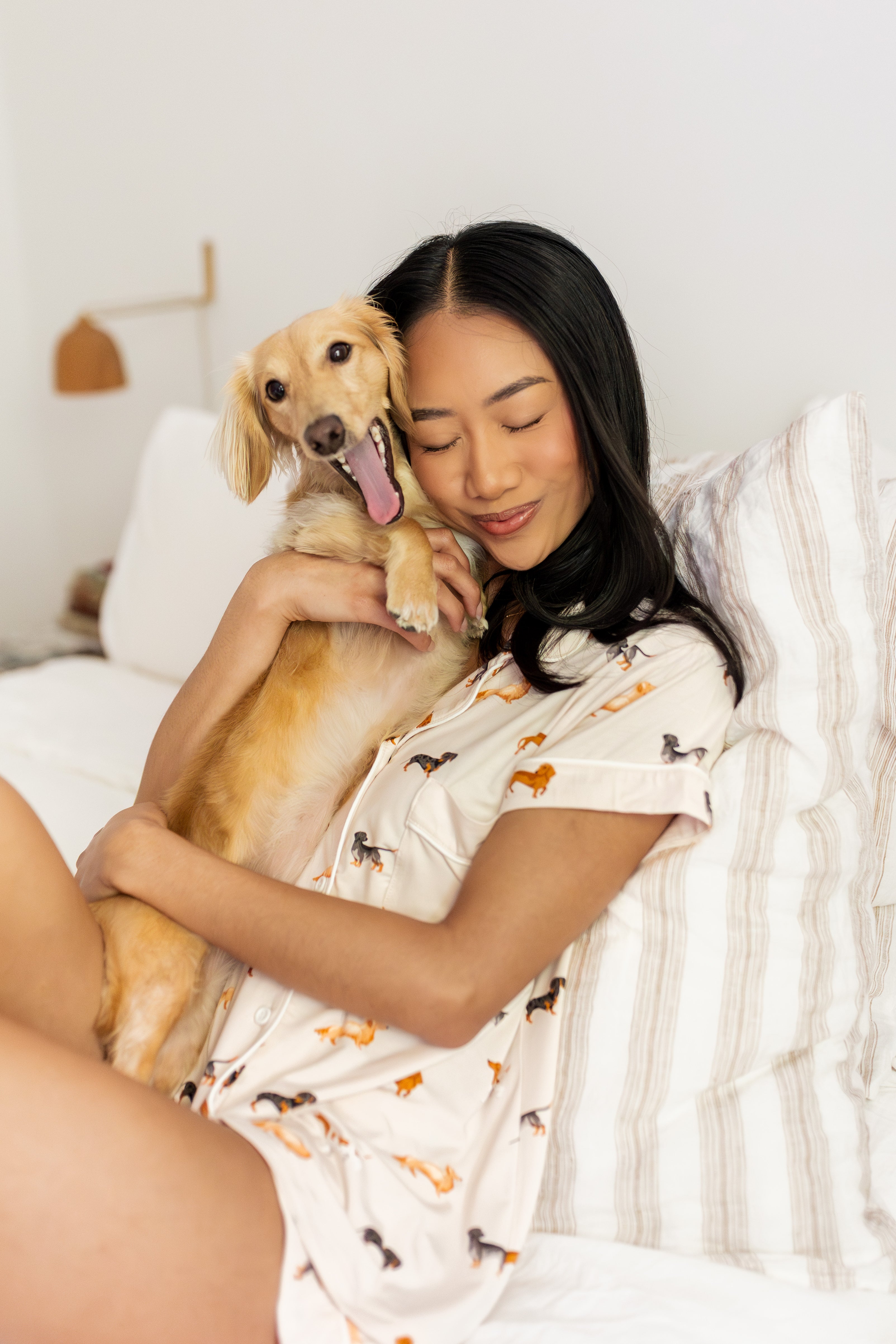 Woman holding blonde yawning dachshund while sitting on a white bed and modeling the Short Sleeve Pajama Set in Dachshund print 
::: Model is 5'6" wearing size XS ///

---PJ 4