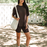 Woman in brown outfit with white sneakers holding a white bag outdoors.