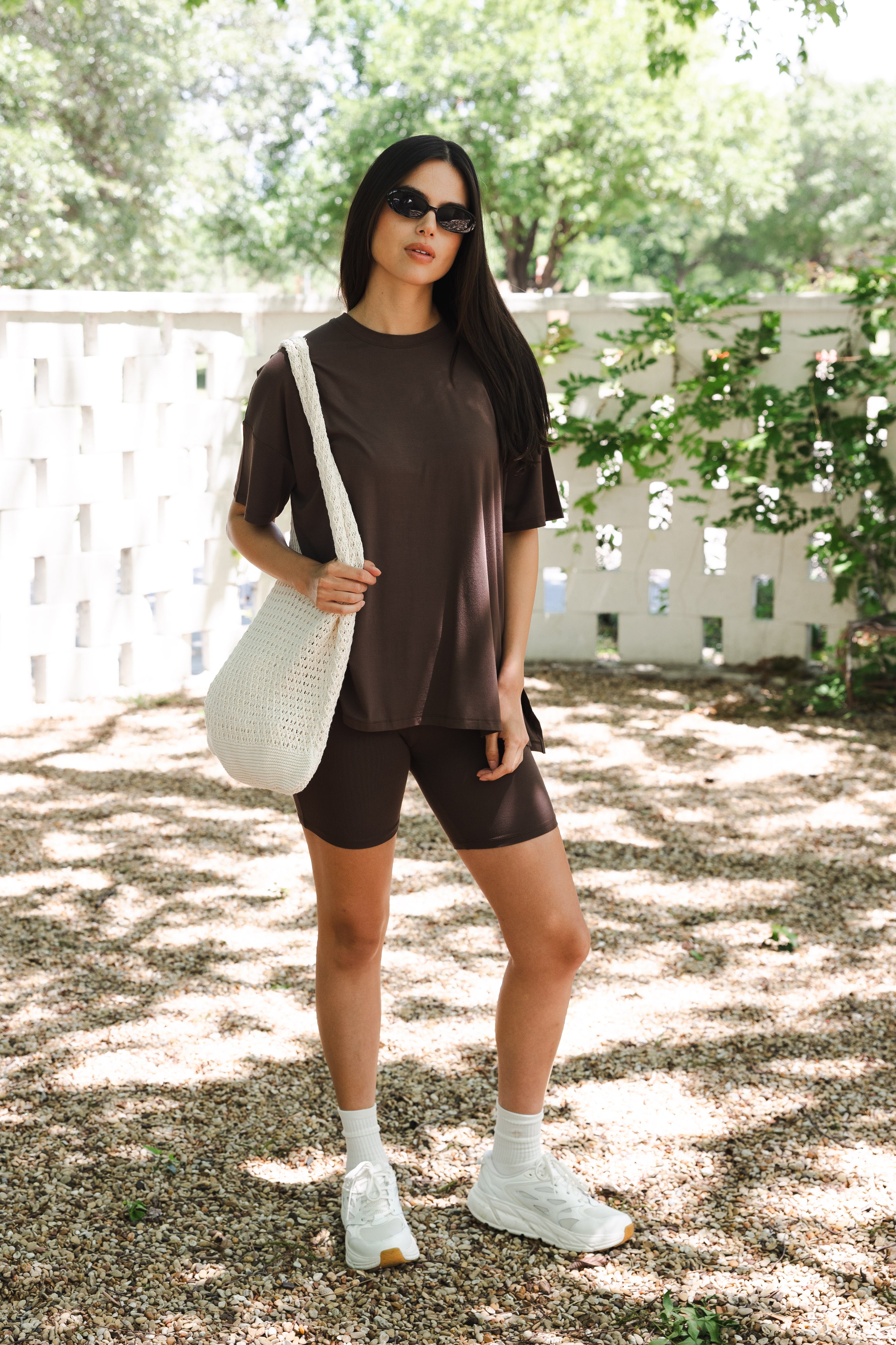 Woman in brown outfit with white sneakers holding a white bag outdoors.
