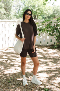 Woman in brown outfit with white sneakers holding a white bag outdoors.