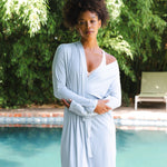 Woman in a light blue robe standing by a pool with greenery in the background
