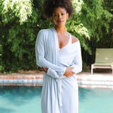 Woman in a light blue robe standing by a pool with greenery in the background