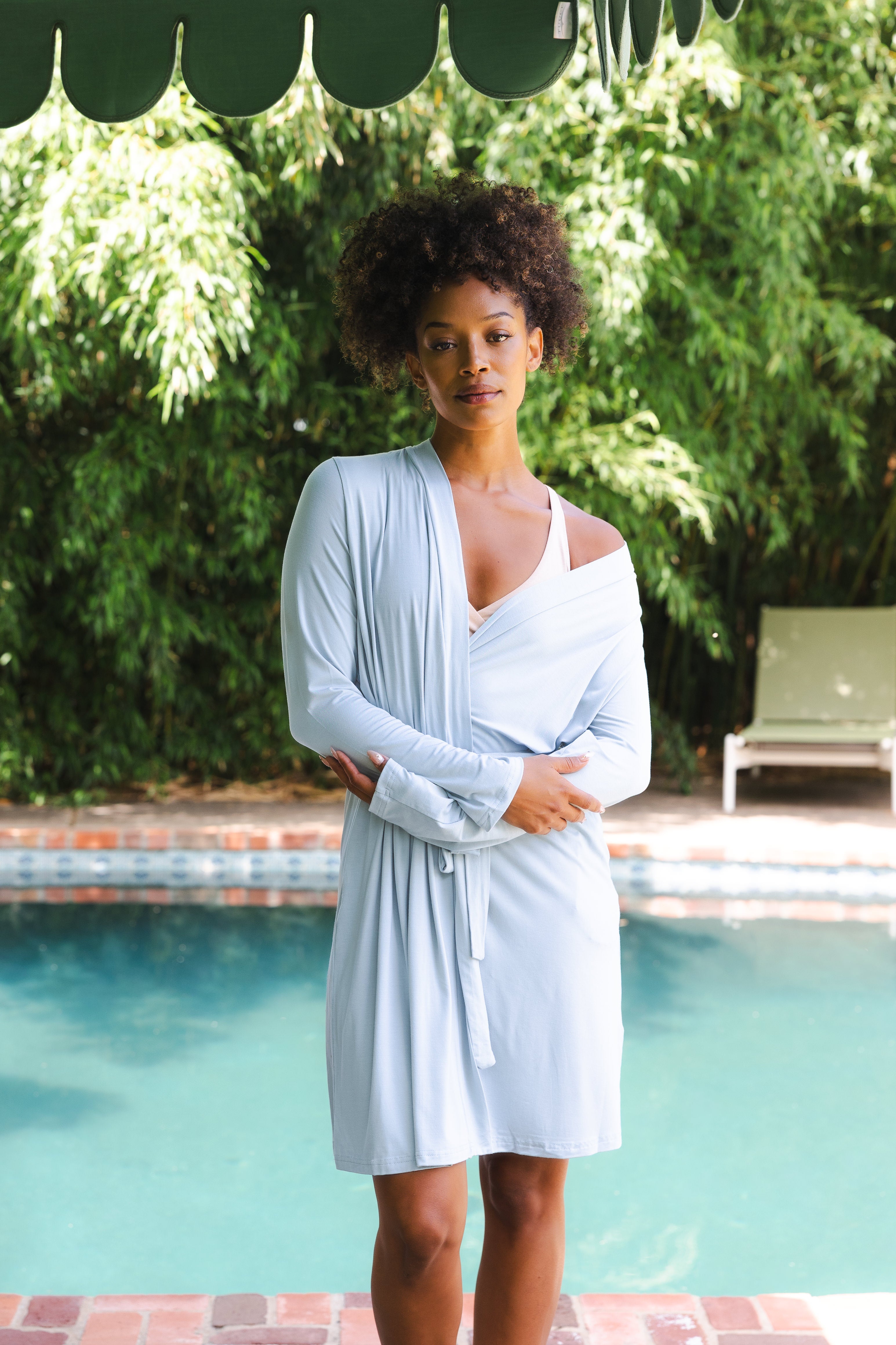 Woman in a light blue robe standing by a pool with greenery in the background