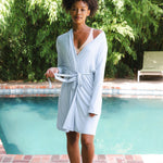 Woman in a light blue robe standing by a pool with greenery in the background