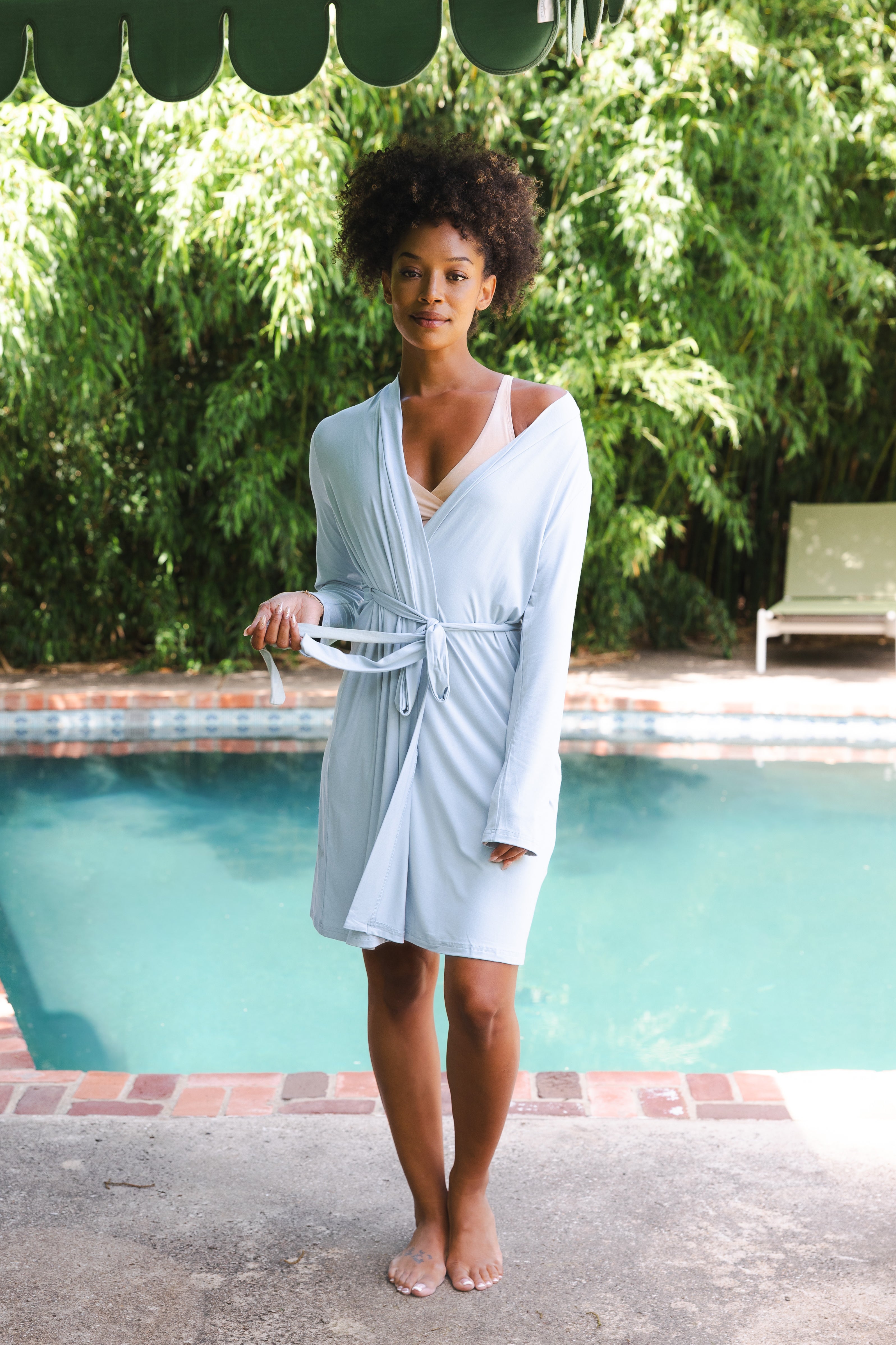 Woman in a light blue robe standing by a pool with greenery in the background