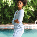 Woman in a light blue robe standing by a pool with greenery in the background
