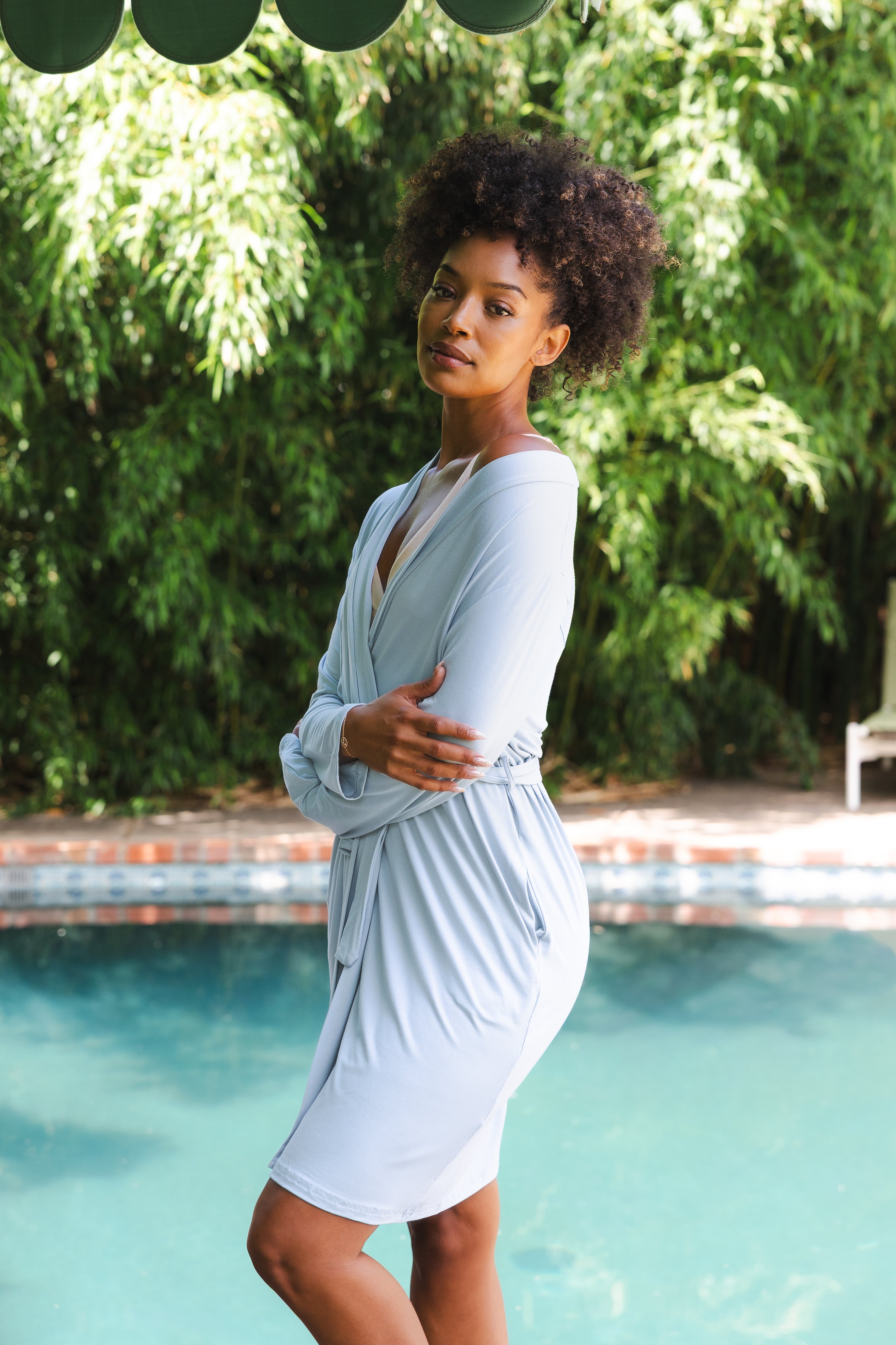 Woman in a light blue robe standing by a pool with greenery in the background