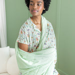 Woman wrapped in a light green blanket with a honeydukes patterned pj against a green wall.
