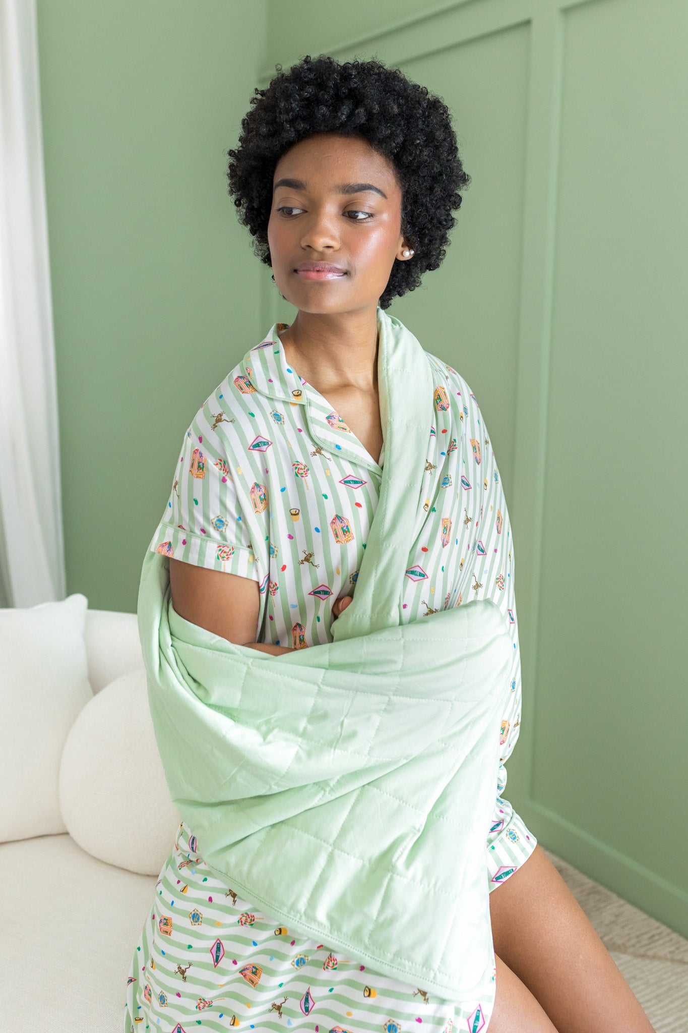 Woman wrapped in a light green blanket with a honeydukes patterned pj against a green wall.
