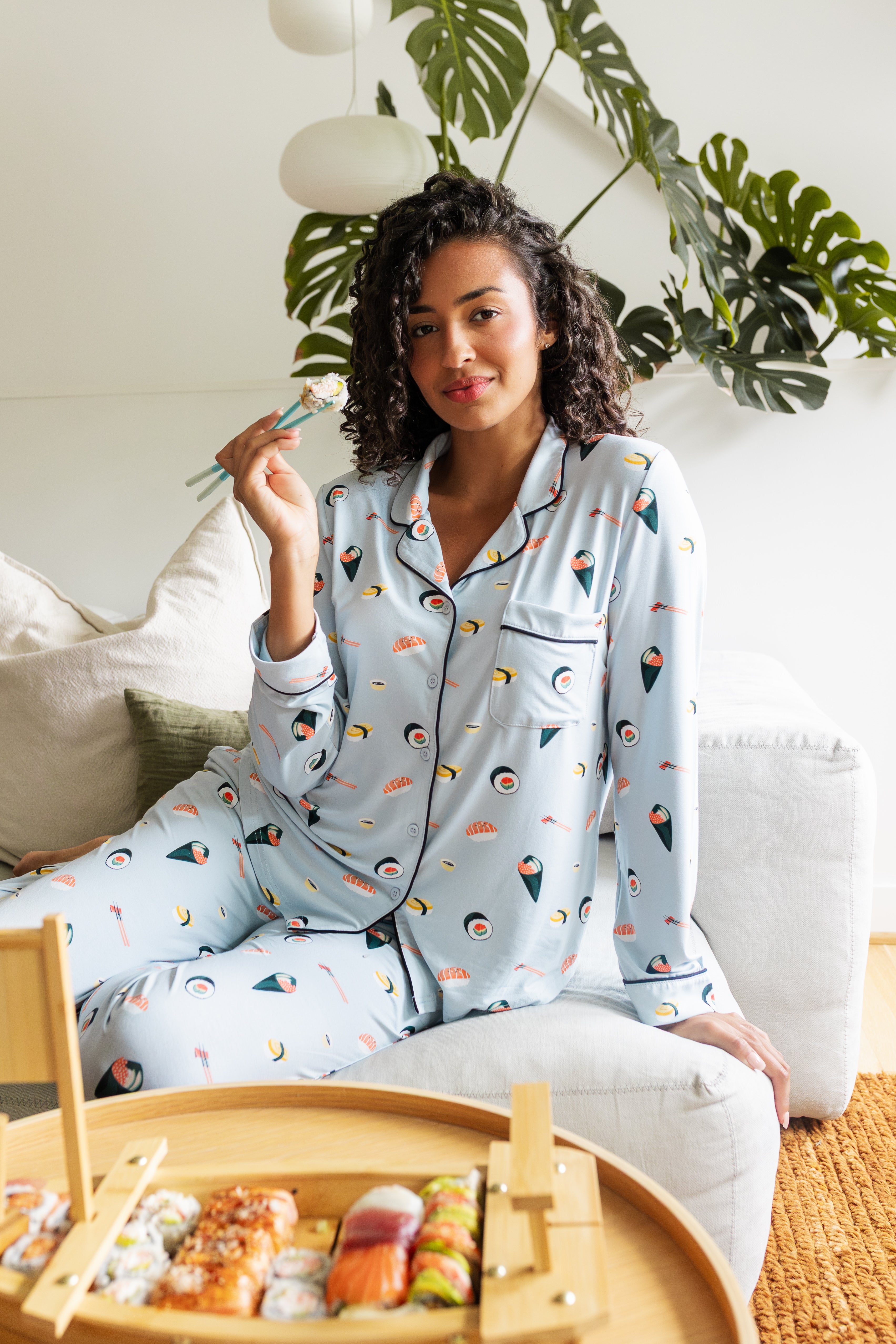Woman wearing a sushi-themed blue pajama set sitting on a couch with a small bamboo basket of sushi.
---1605 1