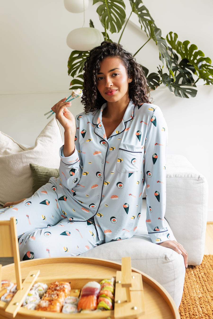 Woman wearing a sushi-themed blue pajama set sitting on a couch with a small bamboo basket of sushi.