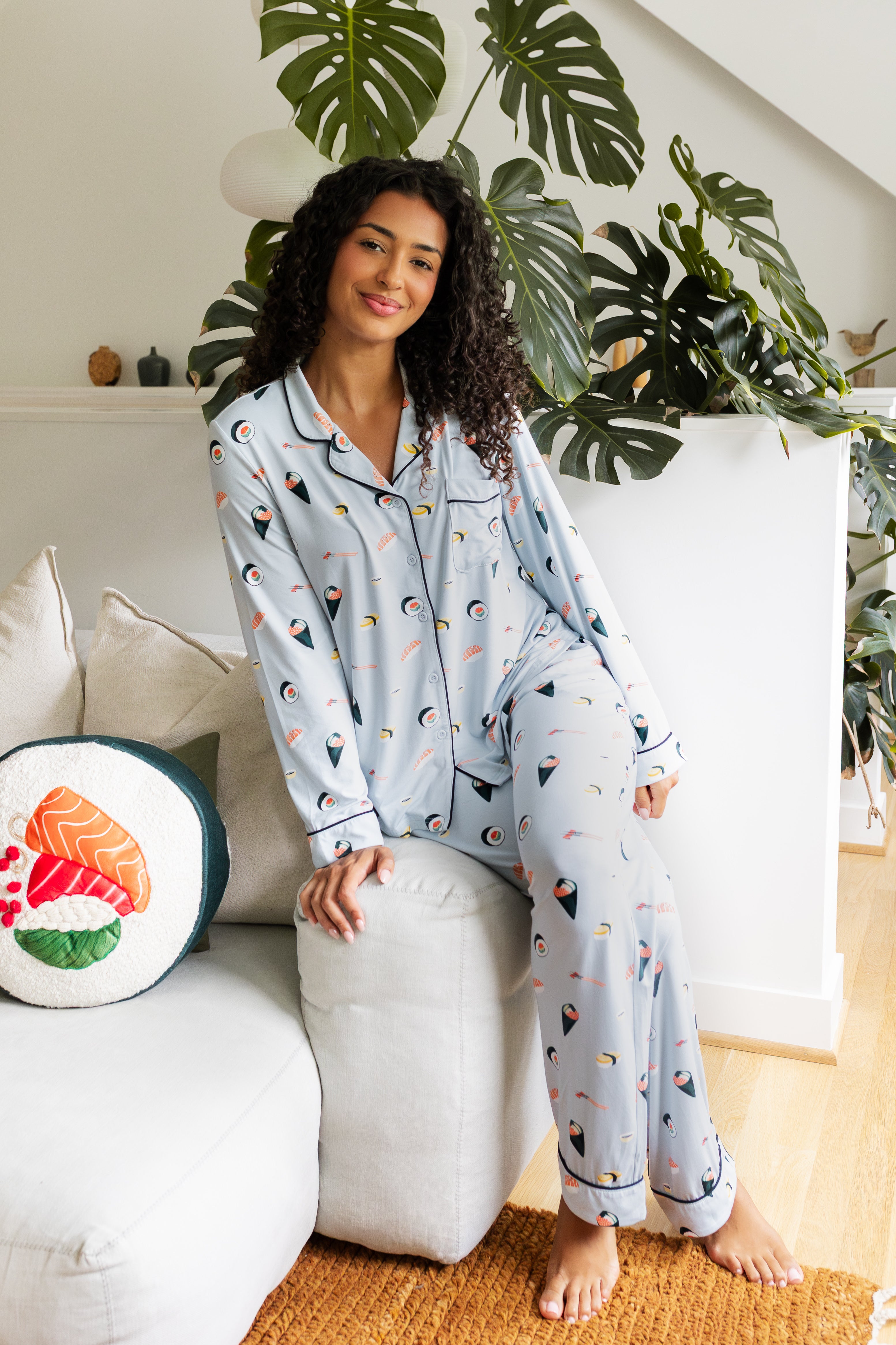 Woman wearing a sushi-themed pajama set sitting on the side of white couch in a cozy living room. ---PJ 4