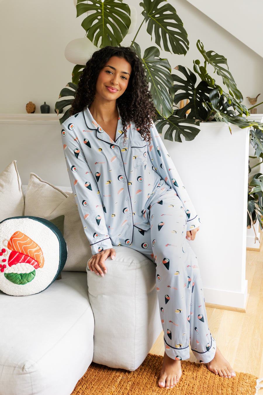 Woman wearing a sushi-themed pajama set sitting on the side of white couch in a cozy living room.