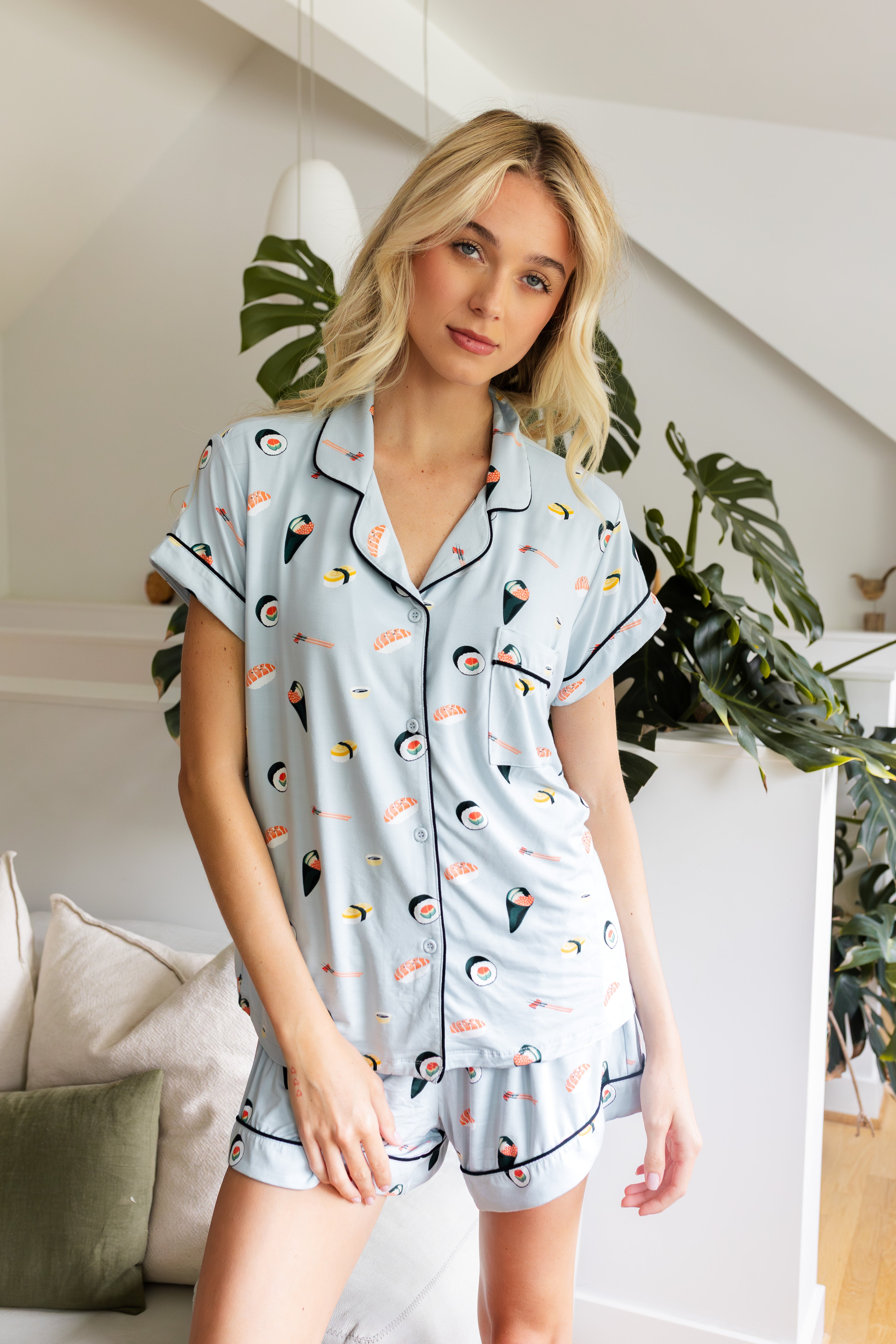 Woman wearing a sushi-themed pajama set in a home setting with plants. 