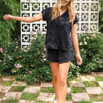 Woman in a black leopard patterned outfit standing outdoors with greenery and a decorative wall in the background
