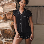 Woman wearing a black pajama set with white trim against a stone wall.