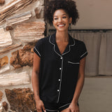 Woman wearing a black pajama set with white buttons standing against a stone wall.