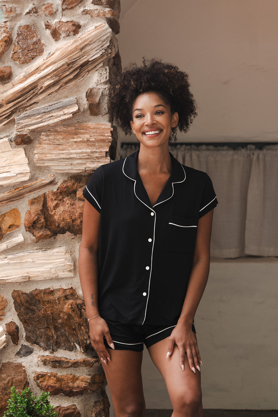 Woman wearing a black pajama set with white buttons standing against a stone wall.