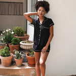 Woman in black pajama set standing outdoors with potted plants.