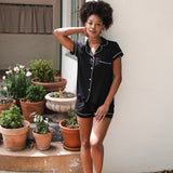 Woman in black pajama set standing outdoors with potted plants.