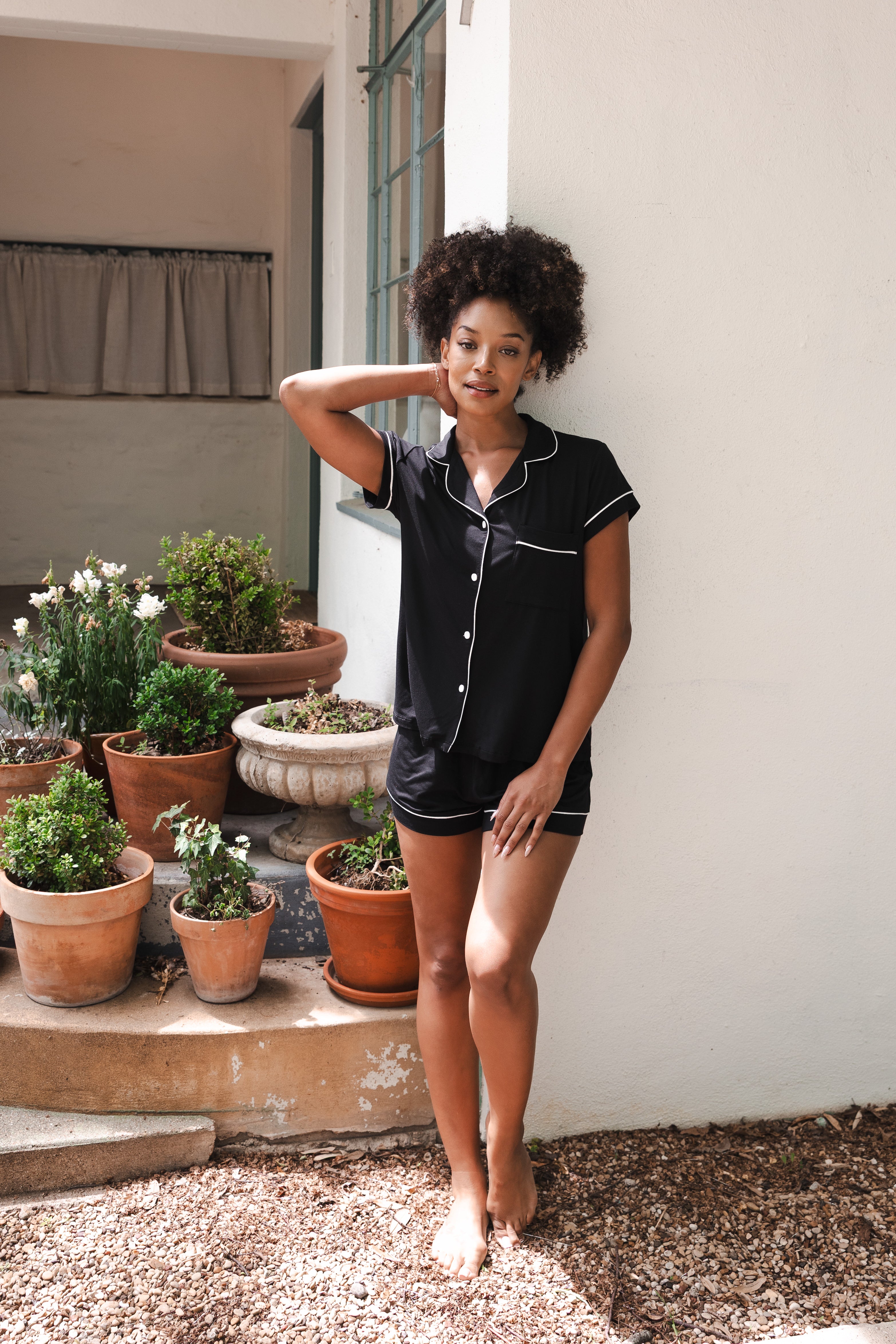 Woman in black pajama set standing outdoors with potted plants.