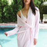 Woman in a light pink robe standing by a pool with greenery in the background