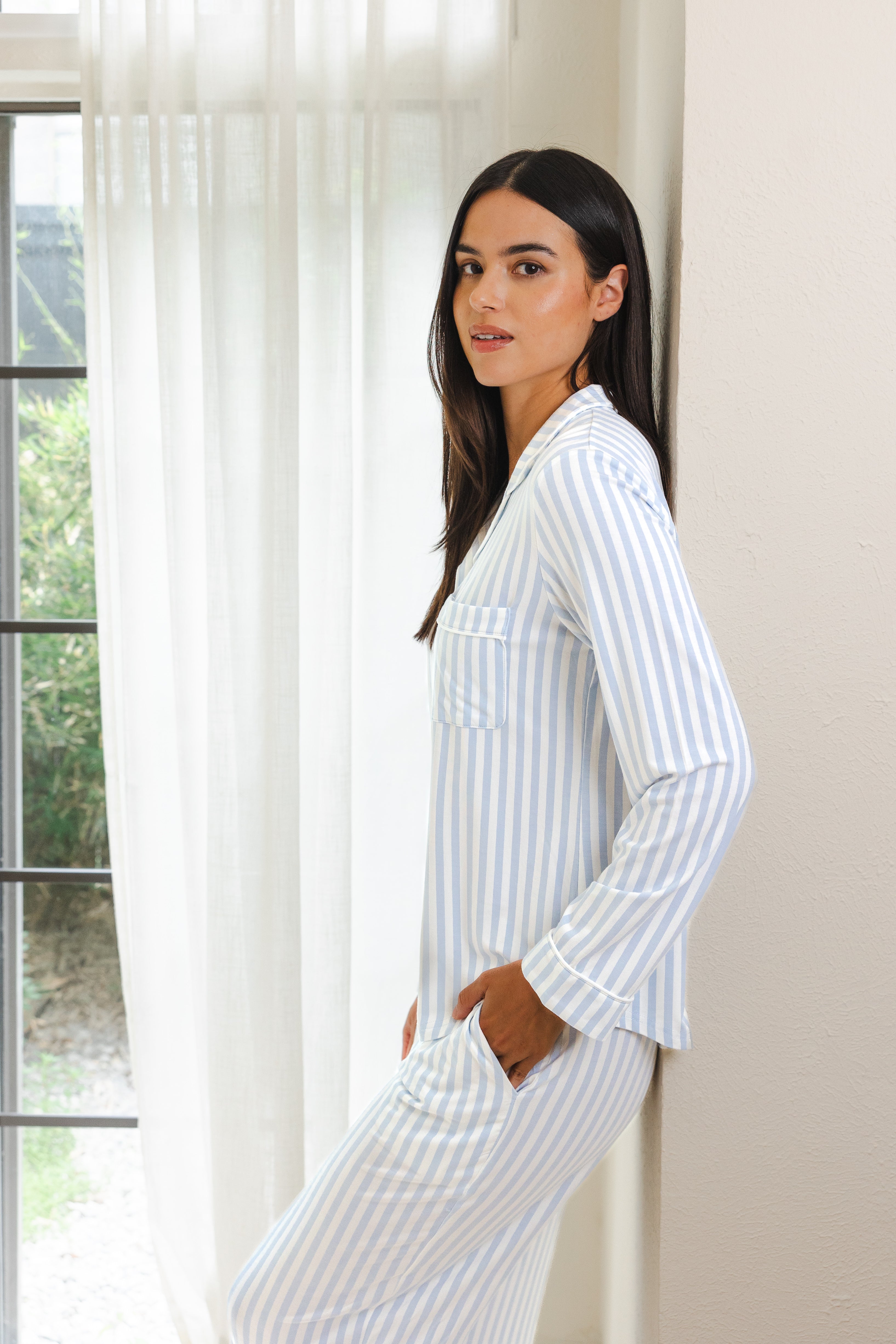 Woman wearing a blue and white striped pajama set standing by a window.