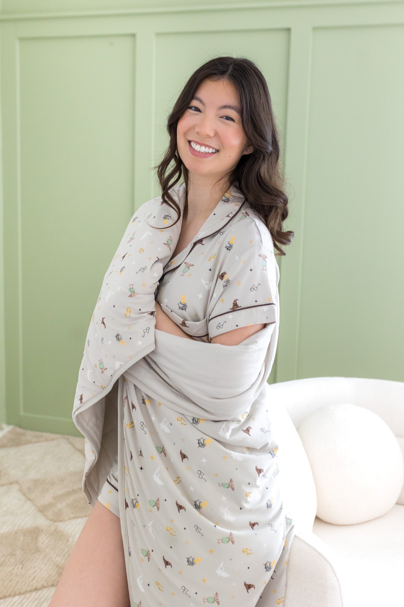Smiling model wrapped in the Throw Blanket in Starry Icon 1.0 wearing matching short sleeve pajama set in front of a light green paneled wal 