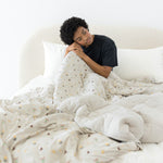 Model sitting on a beige bed covered with the Quilt Blanket in Starry Icon 2.5 with head on knee and wearing crew neck in Midnight 