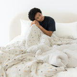 Model sitting on a beige bed covered with the Quilt Blanket in Starry Icon 2.5 with head on knee and wearing crew neck in Midnight 