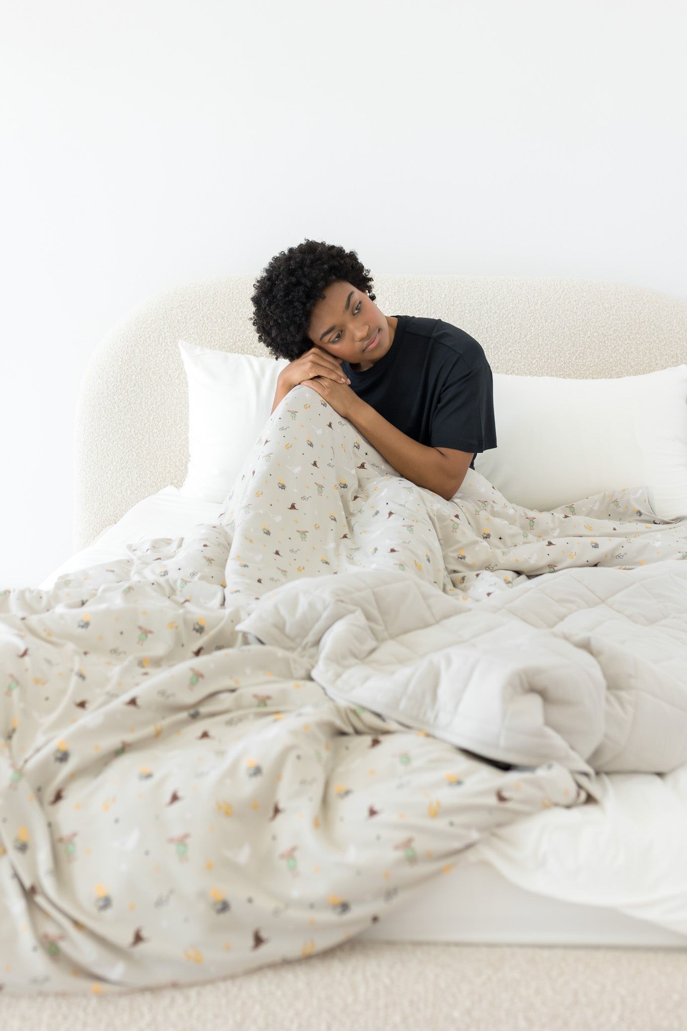 Model sitting on a beige bed covered with the Quilt Blanket in Starry Icon 2.5 with head on knee and wearing crew neck in Midnight 