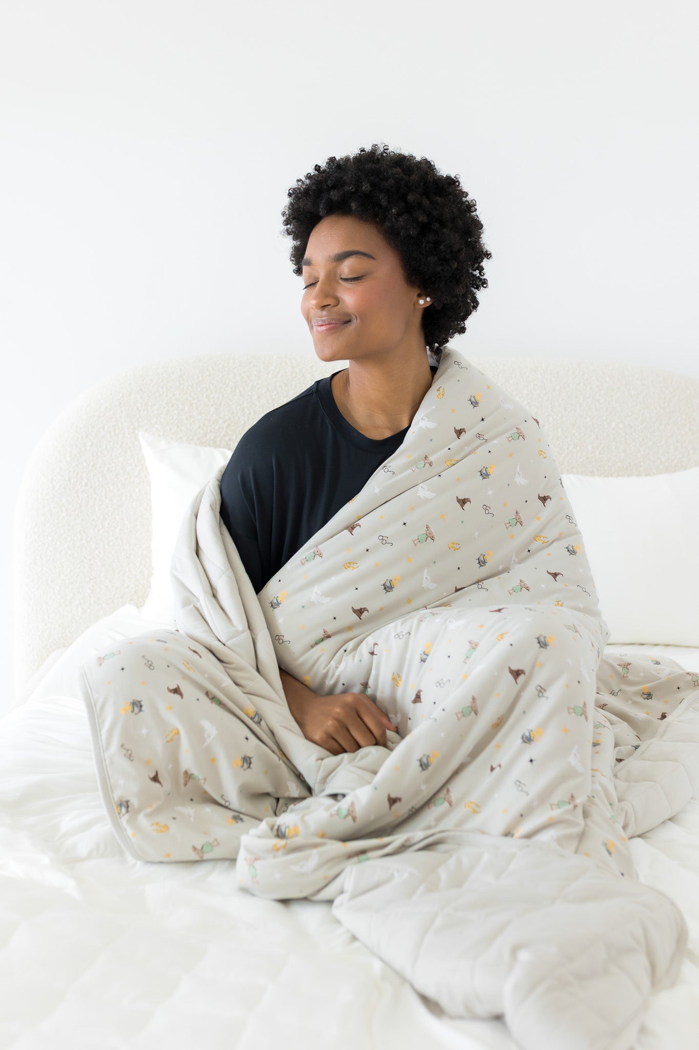 Model sitting on a bed with closed eyes and wrapped in the Quilt Blanket in Starry Icon 2.5
