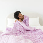Model sitting on a white bed with Adult Blanket in Thistle Leopard print wearing similar print pajamas