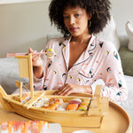 Woman wearing pink Long-Sleeved Women's Pajama Set in Sushi print while eating a meal of sushi in a bamboo tray.