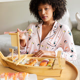 Woman wearing pink Long-Sleeved Women's Pajama Set in Sushi print while eating a meal of sushi in a bamboo tray.