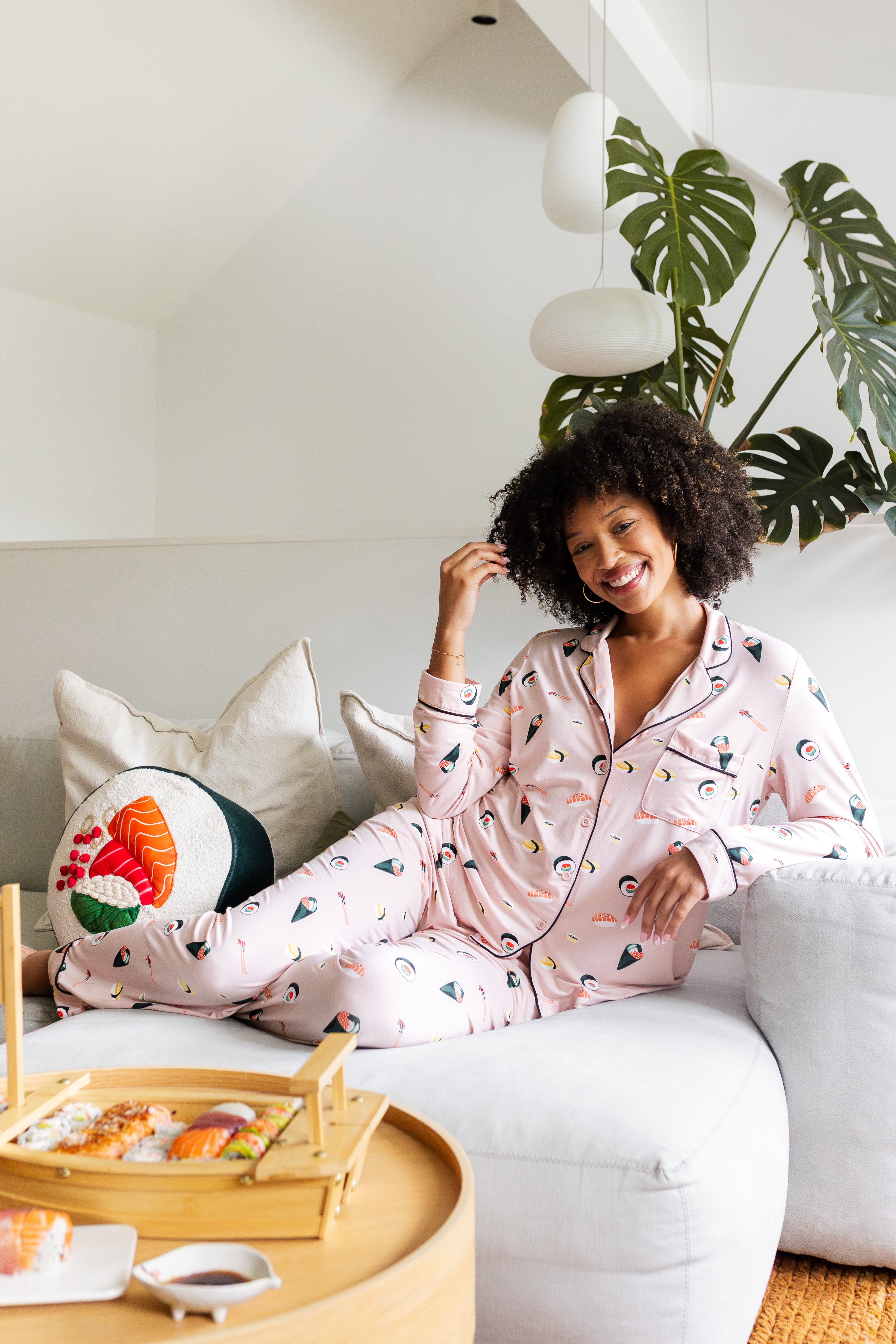 Woman smiling in pink sushi pajamas sitting on a white couch with a plant in the background
