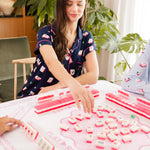Woman wearing mahjong printed pajamas playing a mahjong game with pink tiles in a cozy room.