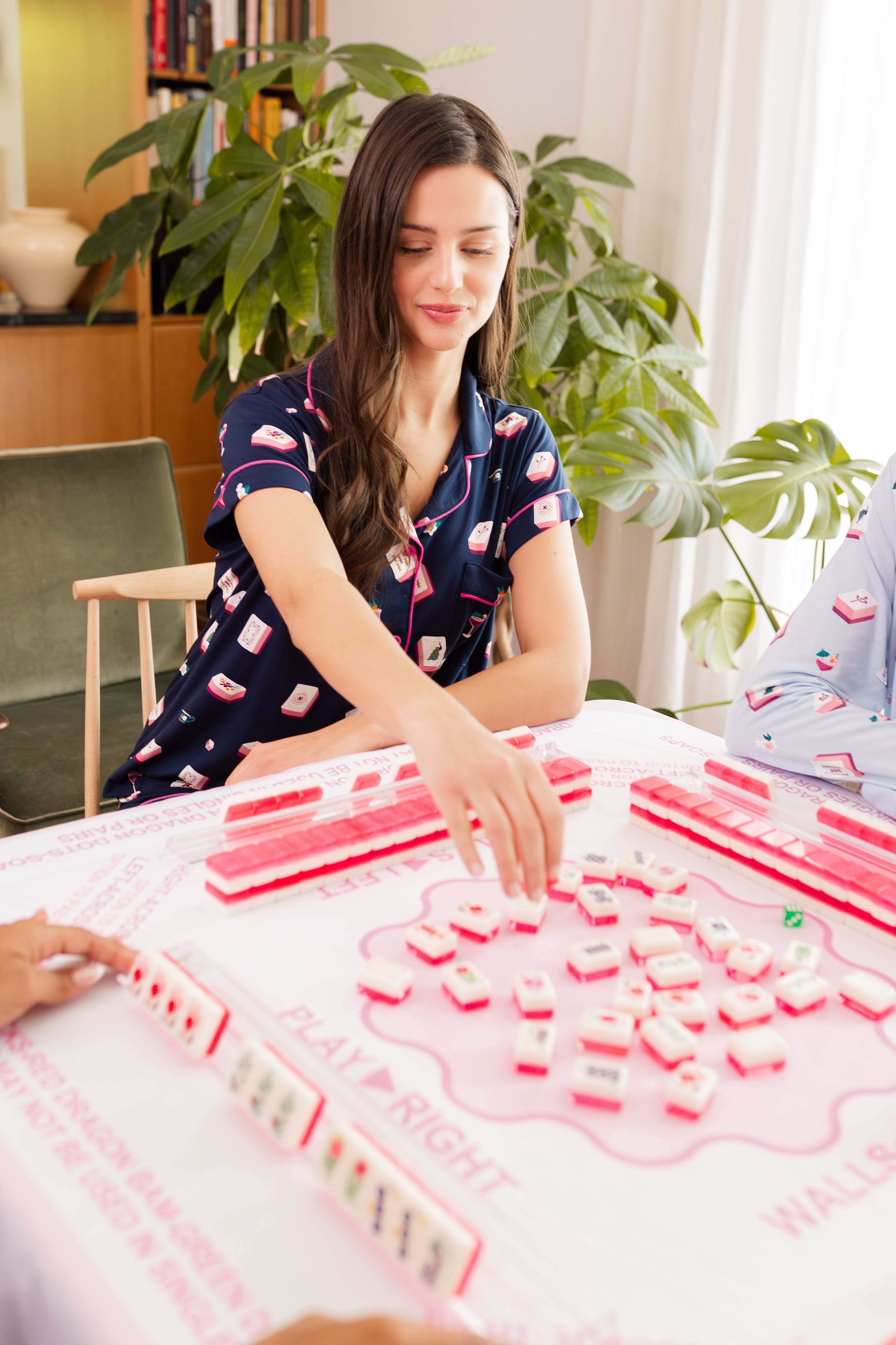 Woman wearing mahjong printed pajamas playing a mahjong game with pink tiles in a cozy room.