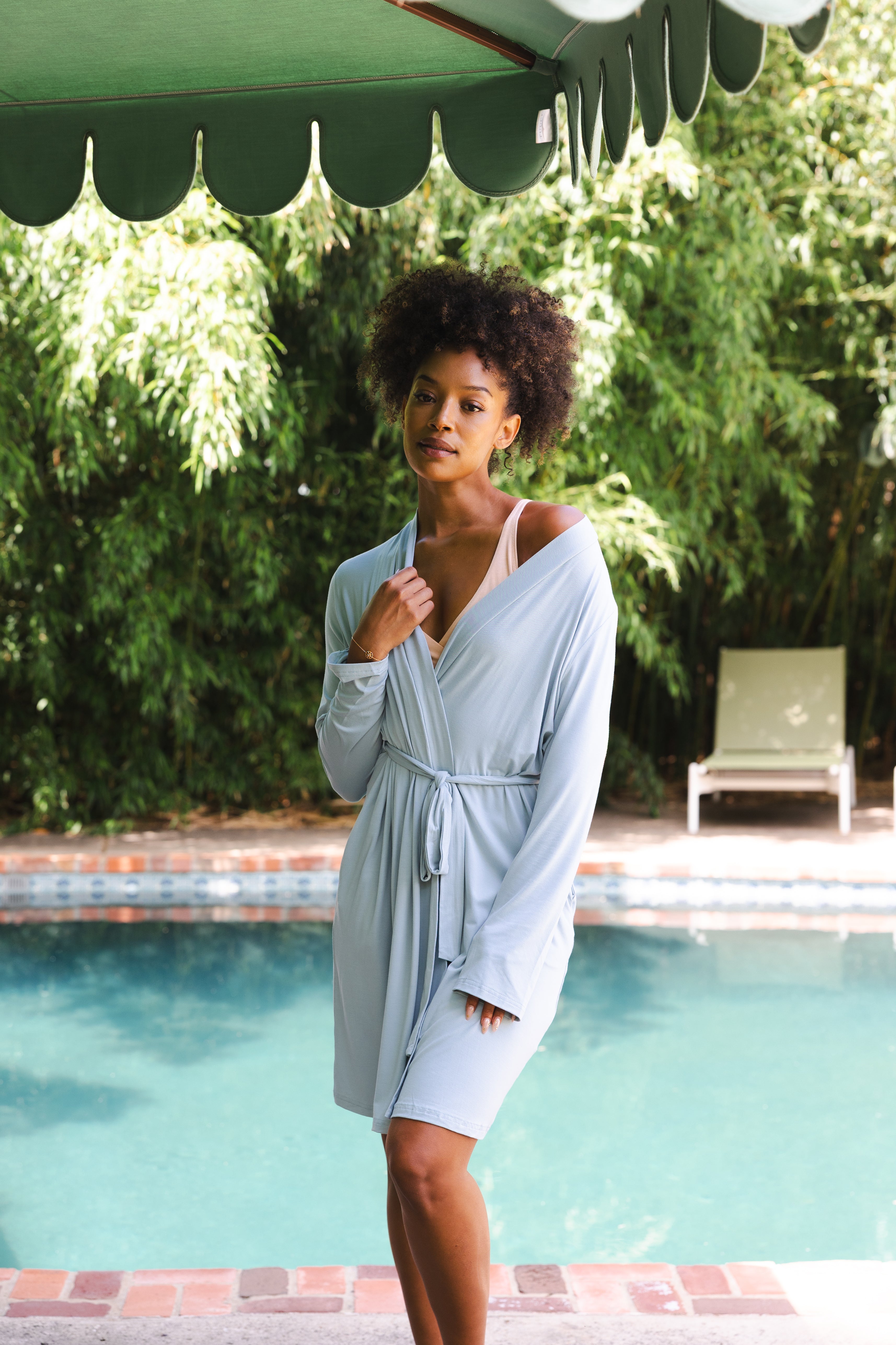 Woman in a light blue robe standing by a pool with greenery in the background