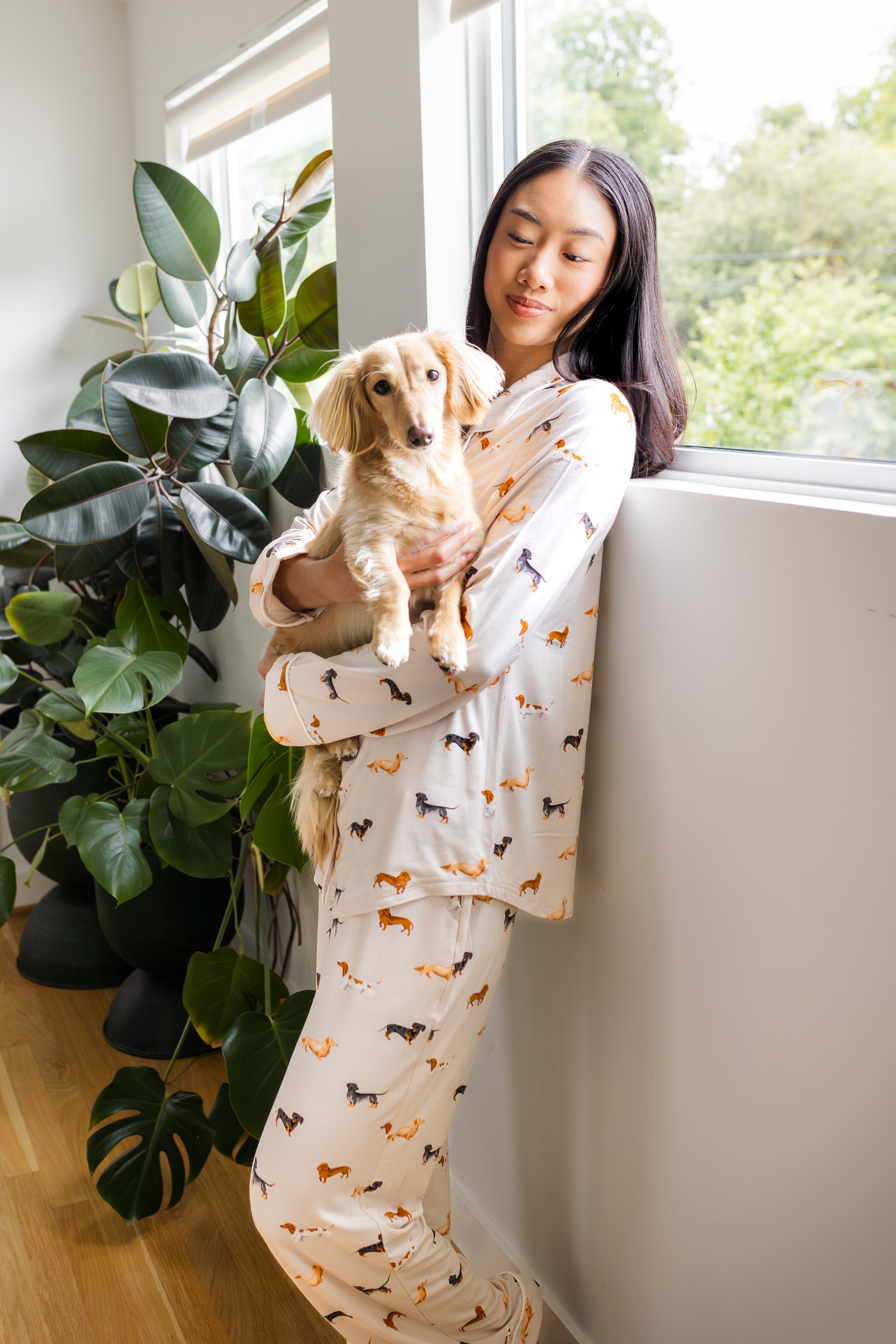 Model wearing Long-Sleeved Women's Pajama Set in Dachshund standing against a wall near a tree holding a Dachshund dog
