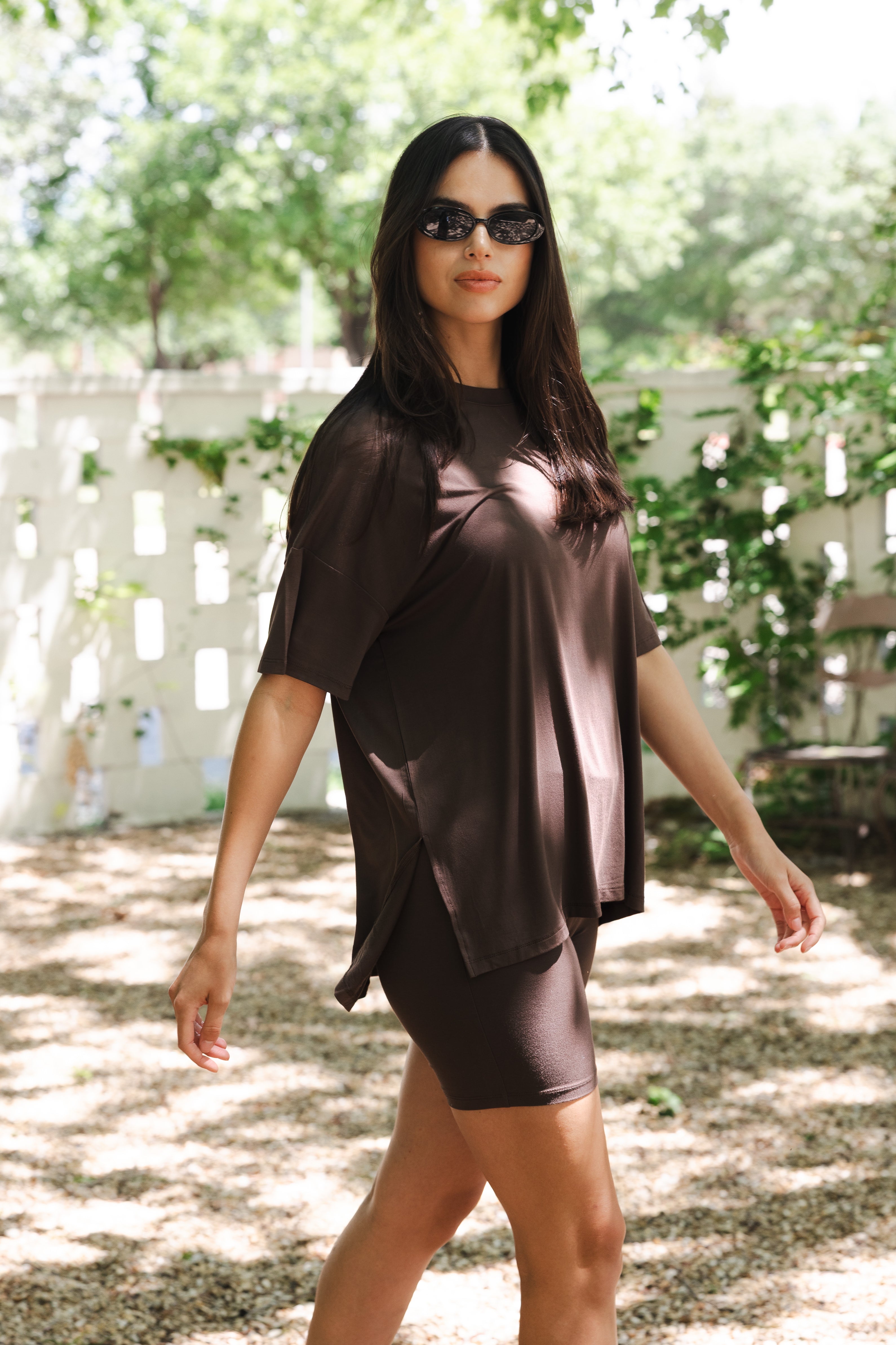 Woman wearing a brown dress and sunglasses outdoors with greenery in the background
