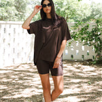 Woman in brown outfit standing outdoors with greenery in the background