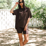 Woman in brown outfit standing outdoors with greenery in the background
