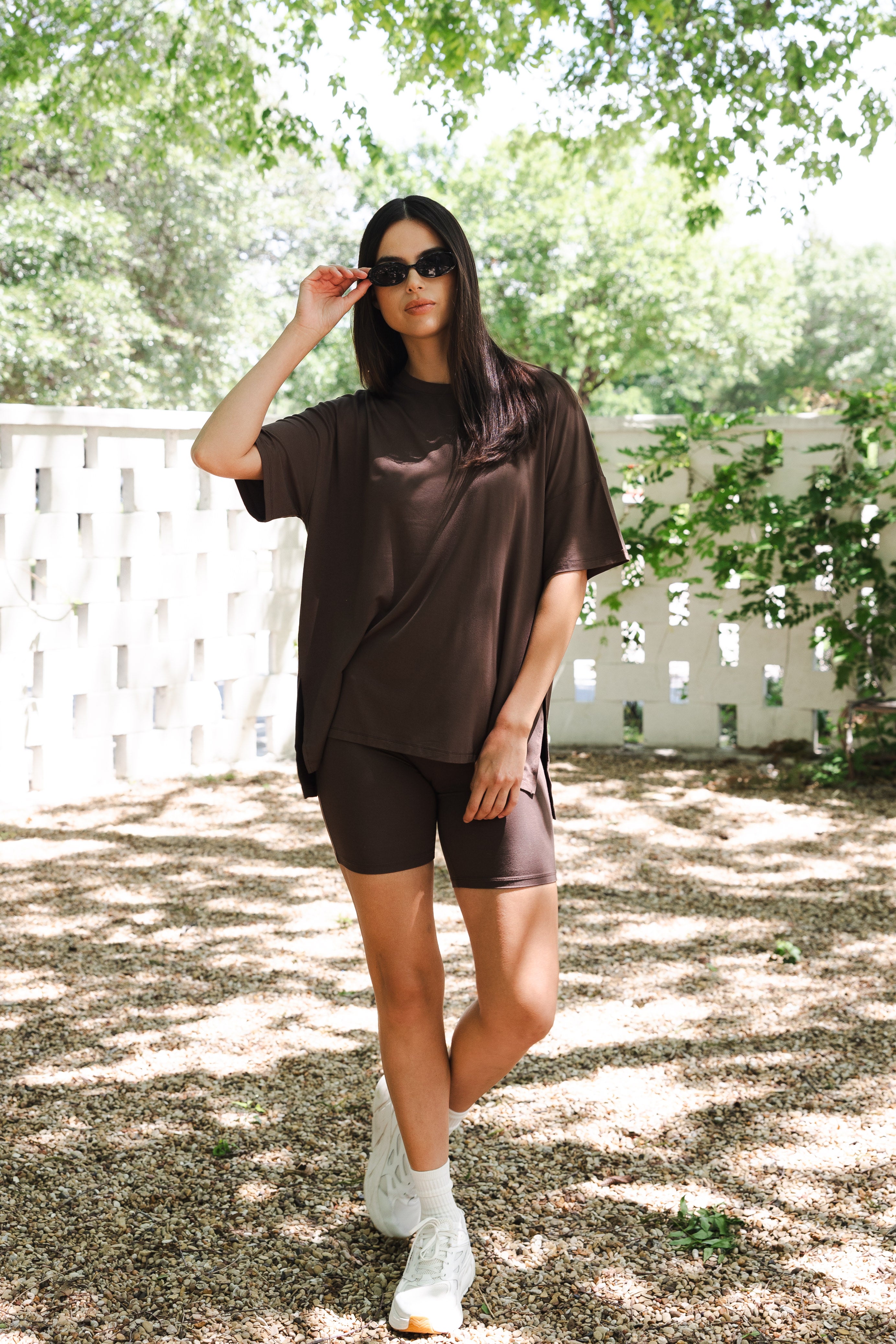 Woman in brown outfit standing outdoors with greenery in the background