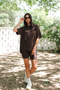 Woman in brown outfit standing outdoors with greenery in the background