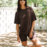 Woman in brown oversized t-shirt and biker shorts standing outdoors with a white fence and trees in the background. ::: Model is 5'10" wearing size S ///