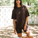Woman in brown oversized t-shirt and biker shorts standing outdoors with a white fence and trees in the background. ::: Model is 5'10" wearing size S ///