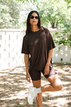 Woman in brown oversized t-shirt and biker shorts standing outdoors with a white fence and trees in the background. ::: Model is 5'10" wearing size S ///